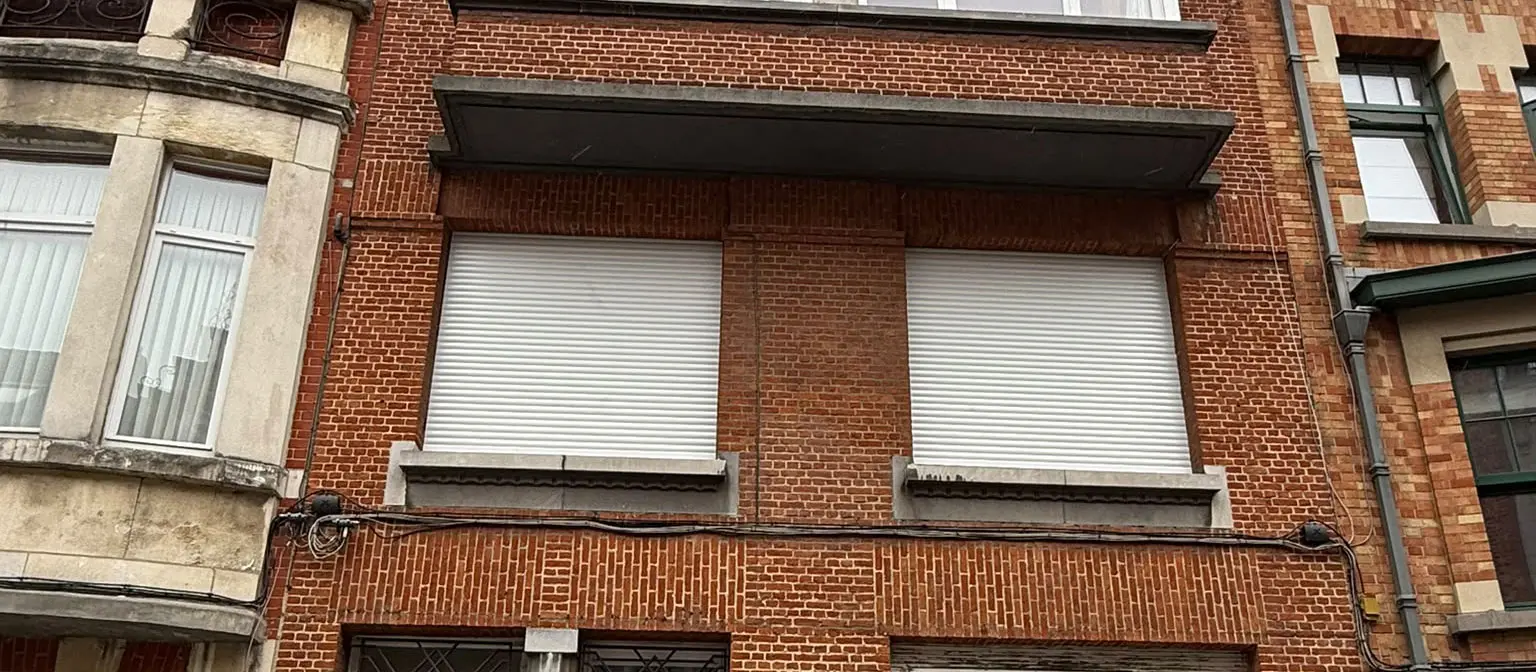 Deux volets aluminium électriques blancs au premier étage d'un immeuble en briques rouges à Schaerbeek, après remplacement des anciens volets bois manuels